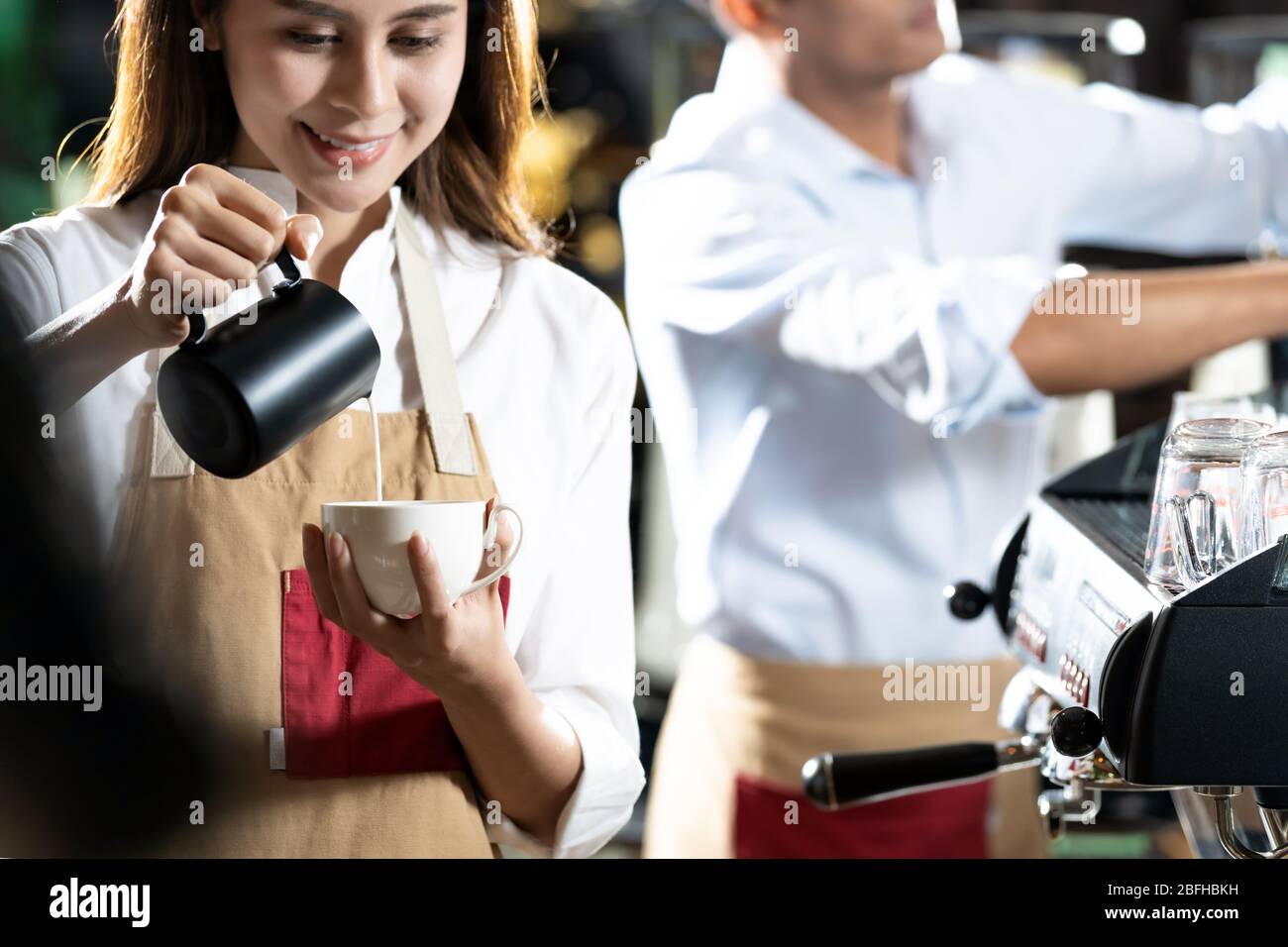 Jeune adulte femme asiatique barista qui verse du lait frais pour préparer du café Latte au client au café-bar avec sa collègue travaillant en arrière-plan. F Banque D'Images