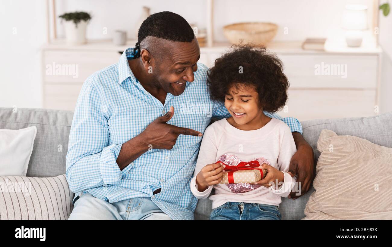 Fête d'anniversaire à la maison. Grand-père afro-américain donnant une boîte cadeau à sa petite-fille à l'intérieur. Panorama Banque D'Images
