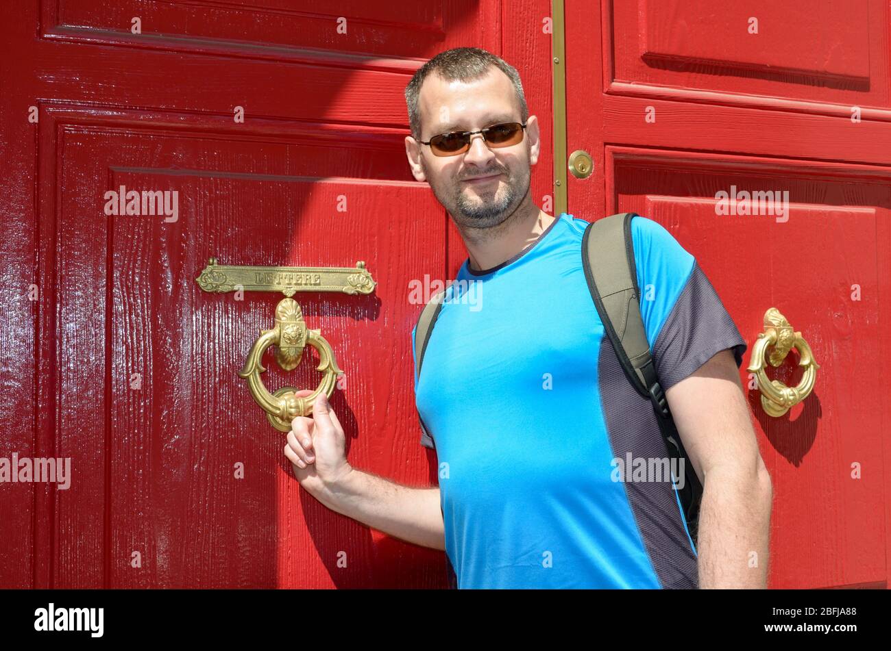 Un touriste d'âge moyen qui frappe à la porte rouge avec un rappeur décoratif de porte et une fente de lettre dorée Banque D'Images