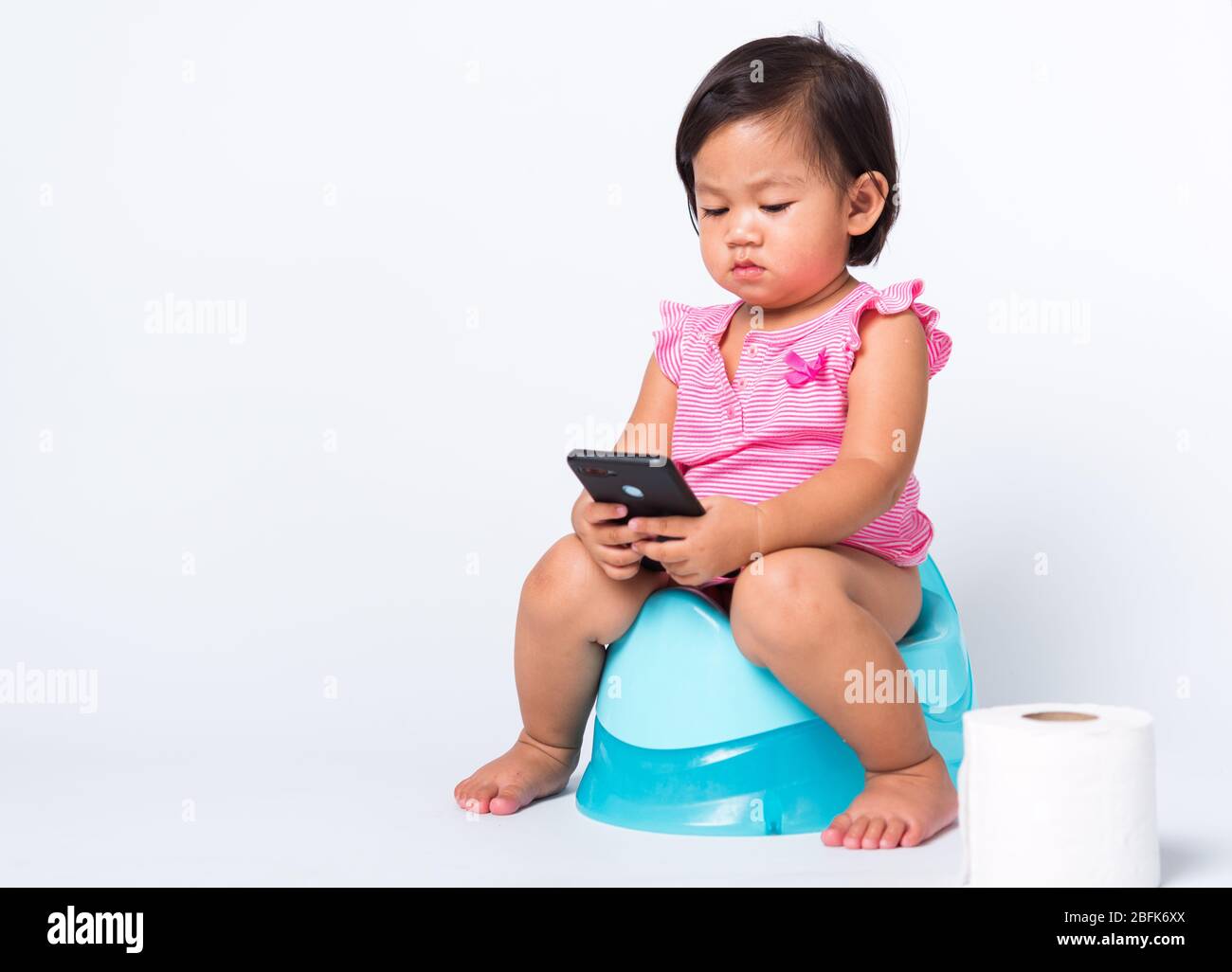 Asiatique petite mignonne bébé fille éducation entraînement à s'asseoir sur pot bleu de chambre ou pot et jouer smartphone mobile avec des rouleaux de papier de toilette, goujon Banque D'Images