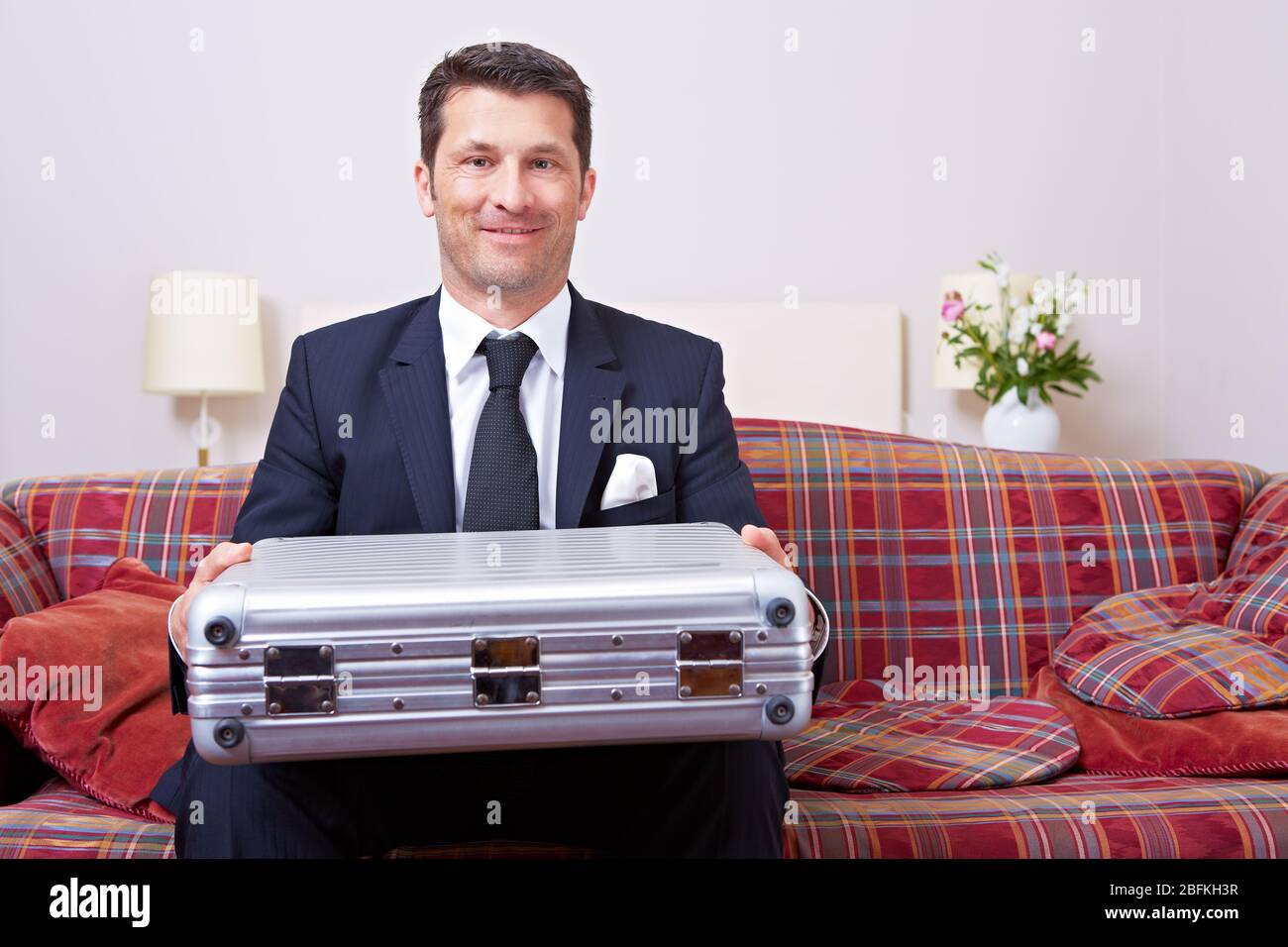 Homme d'affaires souriant assis avec une valise pendant un voyage d'affaires sur un canapé dans l'hôtel Banque D'Images