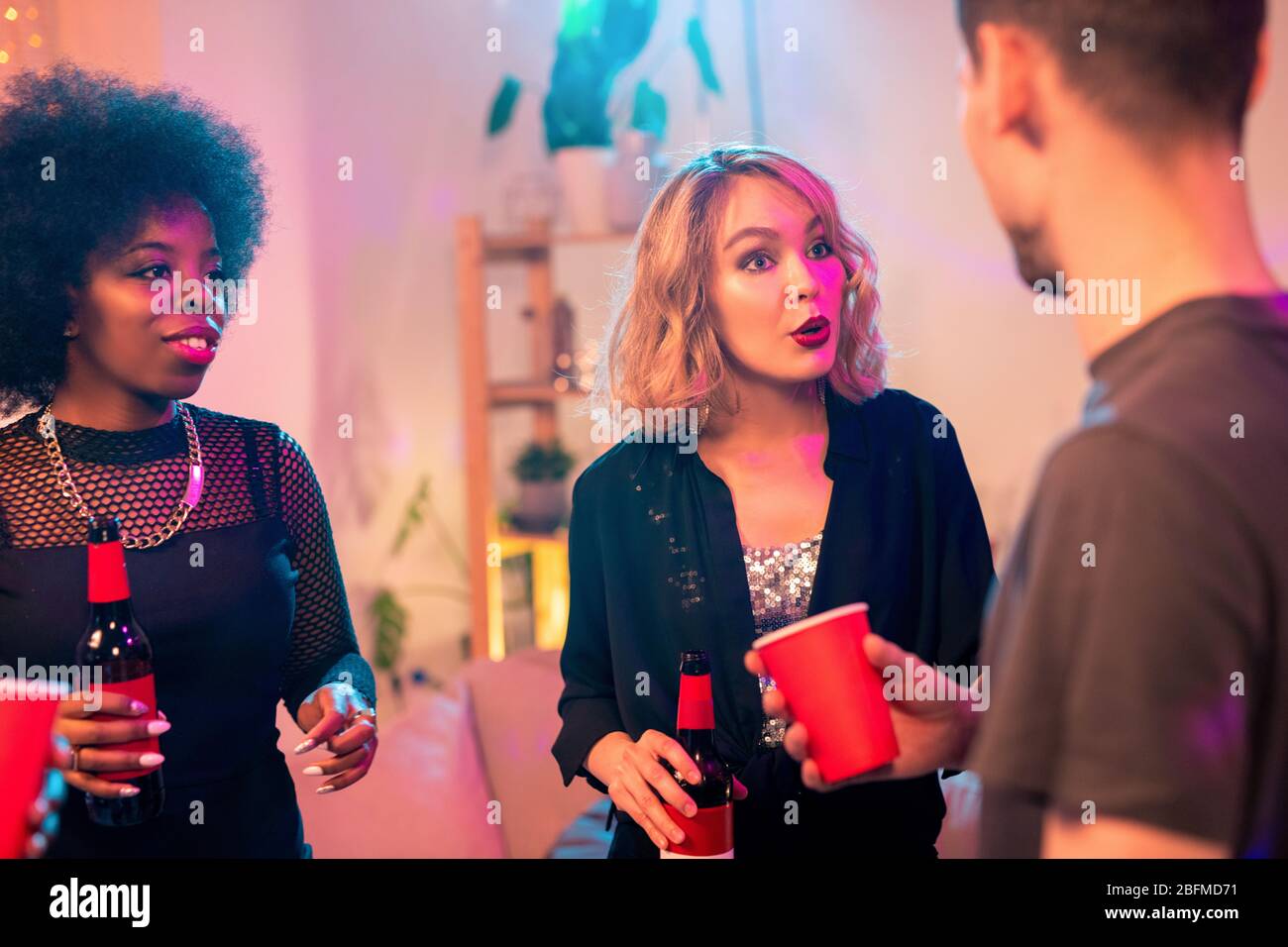Jeune femme blonde avec une bouteille de bière parlant à son ami à la fête tout en prenant des boissons et s'amuser avec des amis Banque D'Images
