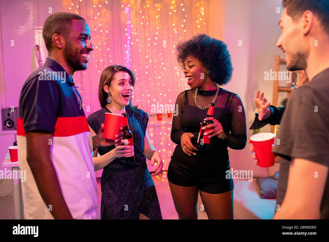 Deux jeunes couples interculturels joyeux dans une tenue glamour avec des boissons à la fête dans l'environnement domestique et discuter de choses drôles Banque D'Images