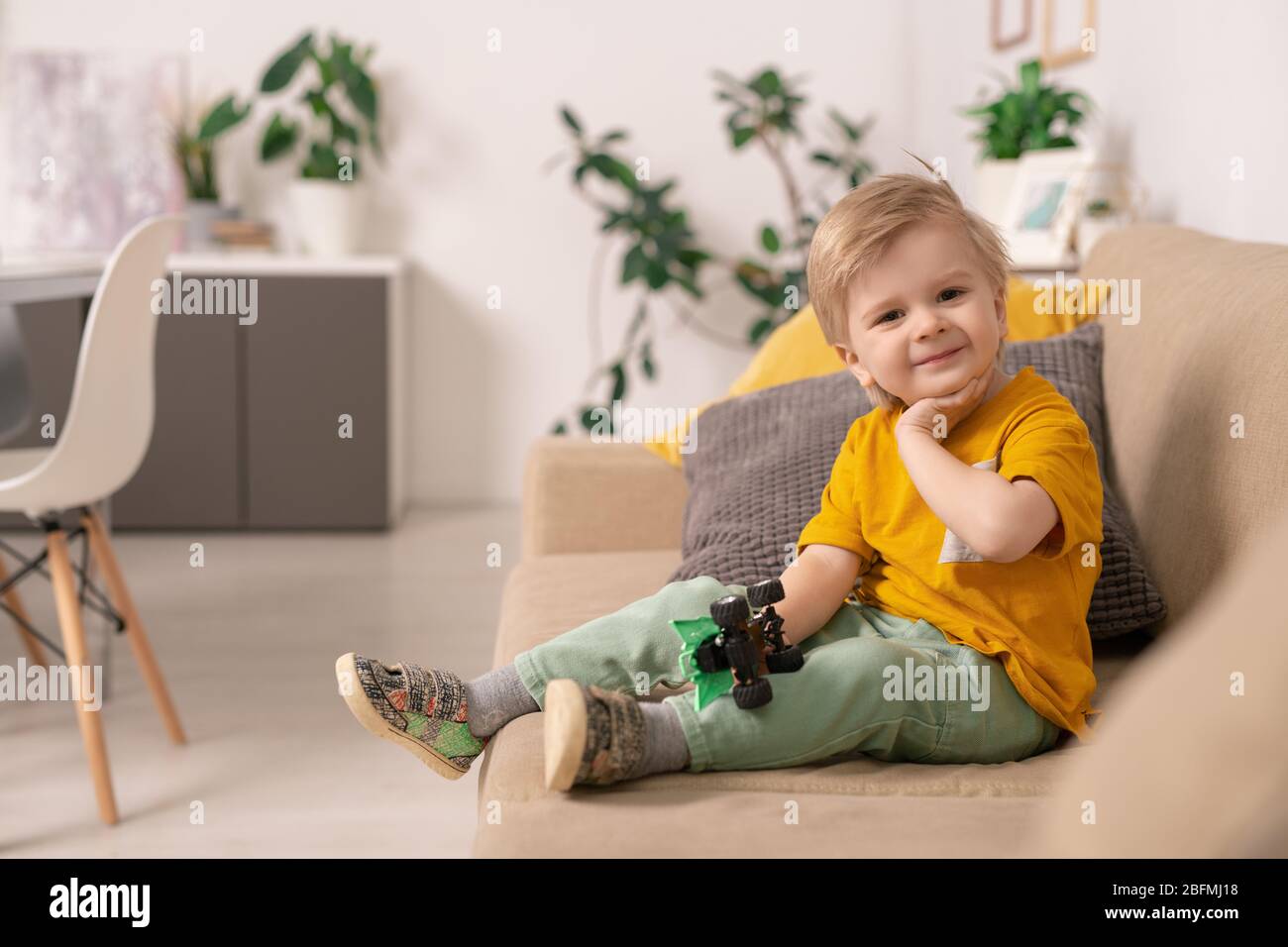 Joyeux joli garçon blond en tenue de bureau assis sur un canapé doux dans le salon et jouer avec une voiture jouet dans l'environnement de la maison Banque D'Images