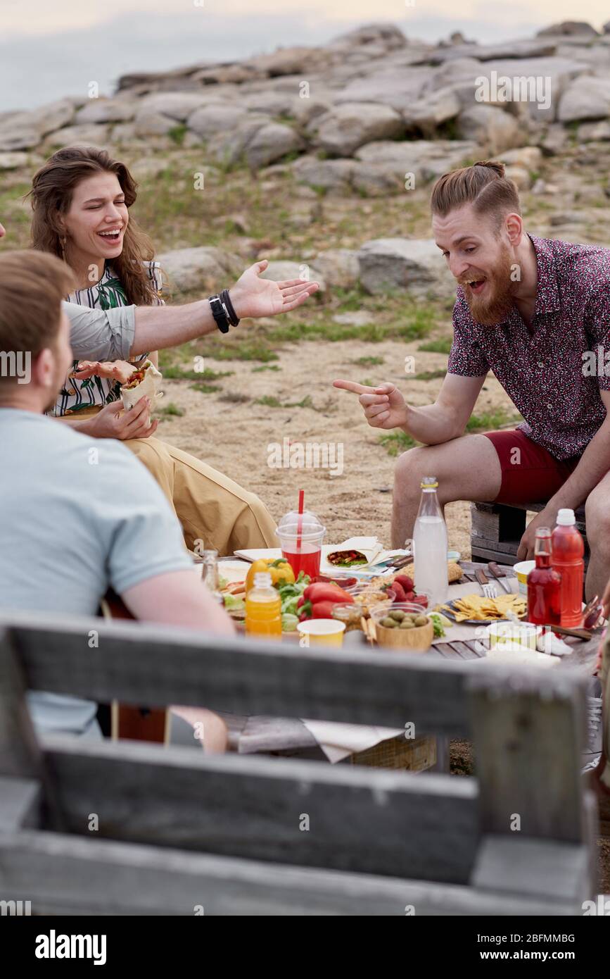 Jeunes Caucasiens assis à table avec nourriture et boissons, bavarder et rire avec joie tout en passant la soirée d'été à l'extérieur Banque D'Images