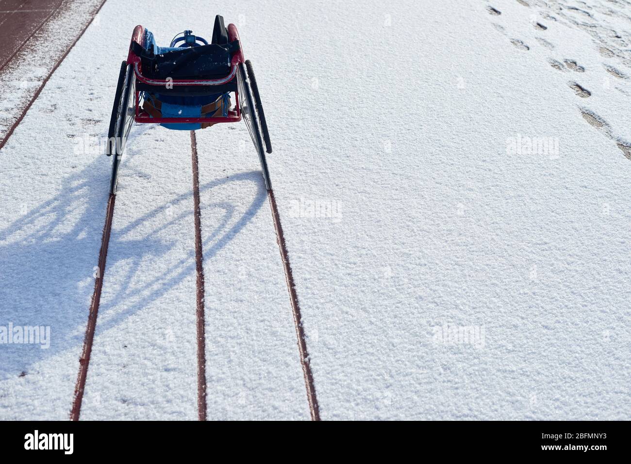 Vue arrière d'un fauteuil roulant de course moderne vide debout sur une piste extérieure et un stade de terrain avec des traces de roues vues sur la neige Banque D'Images