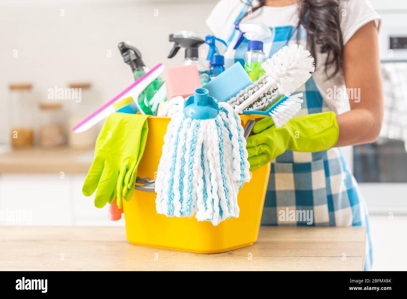 Seau plein de matériel de maintien de maison sur un bureau de cuisine avec une femme le tenant dans des gants en caoutchouc. Banque D'Images