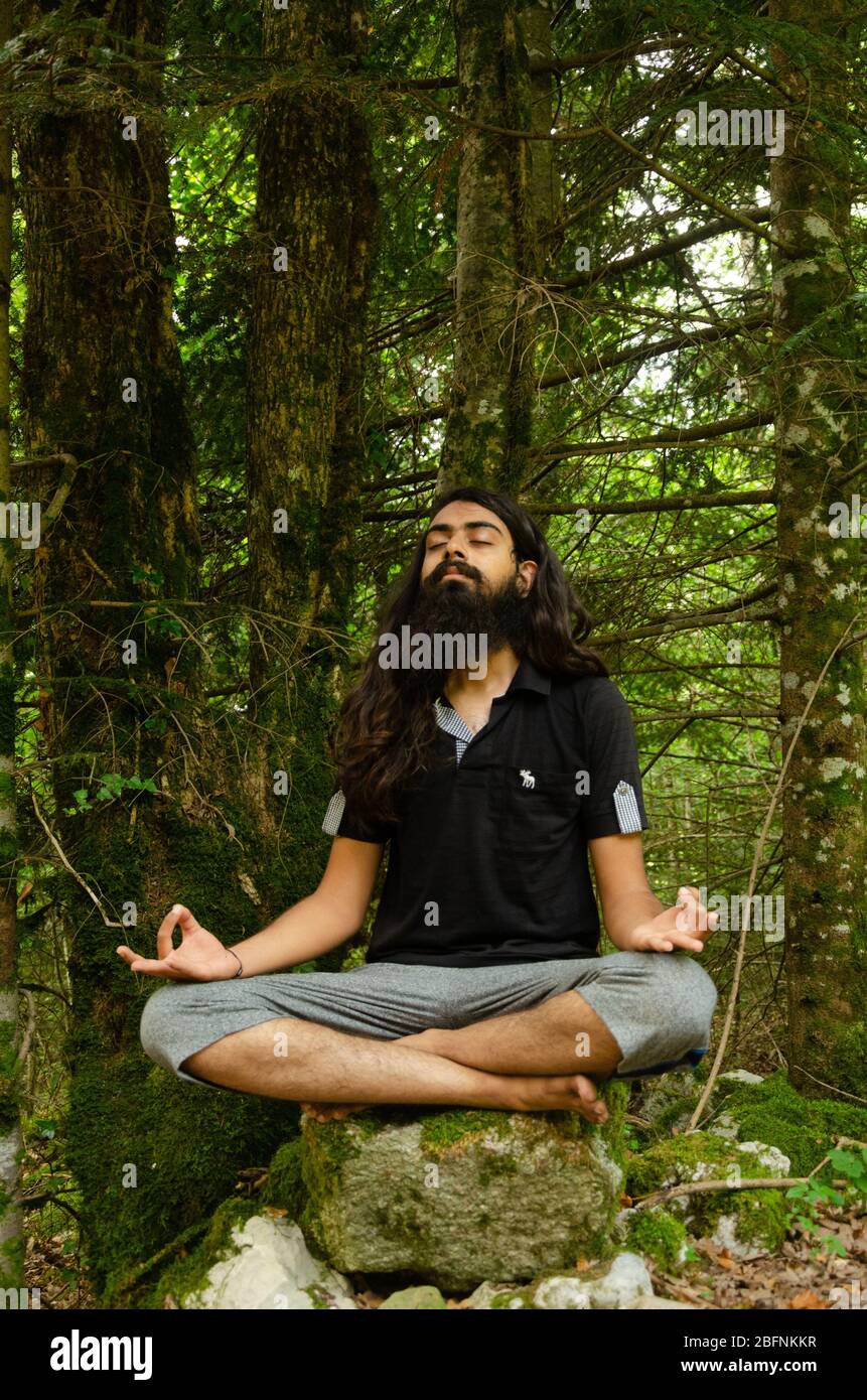 Barbu et cheveux longs garçon indien méditant assis sur un rocher contre la nature forestière toile de fond Banque D'Images