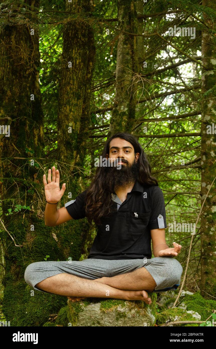 Un garçon indien barbu et à cheveux longs dans la bénédiction pose assis sur une croix sur un rocher contre la nature de la forêt toile de fond Banque D'Images