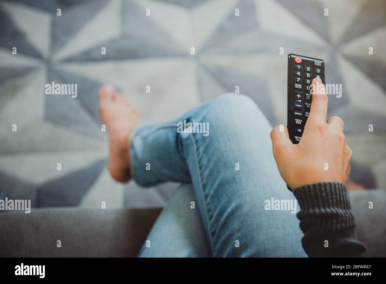 Vue de dessus de la jeune femme tenant la télécommande et allumant le téléviseur tout en étant assise sur un canapé chez elle. Arrêtez le virus Covid-19.Restez à la maison concept. Banque D'Images