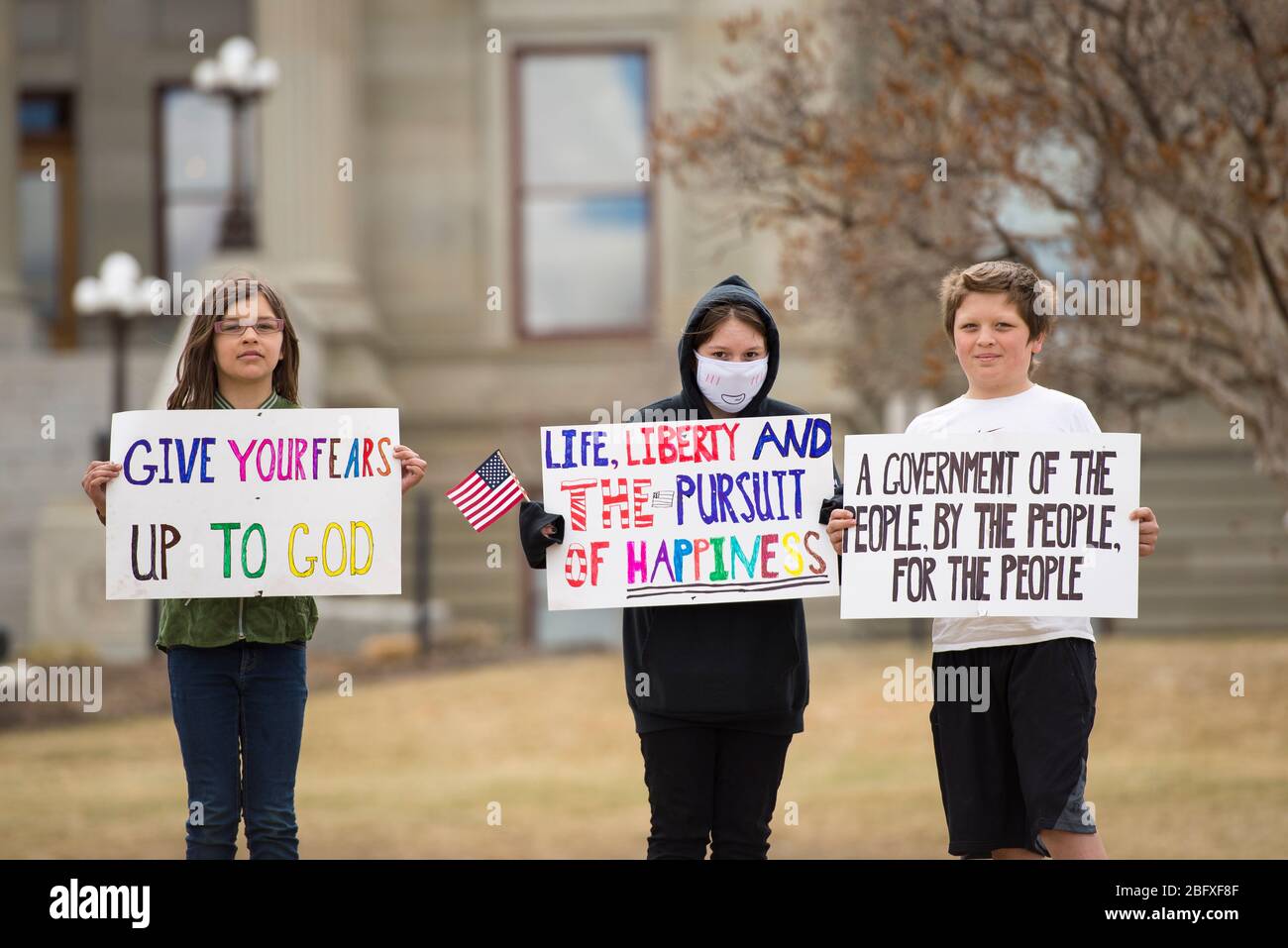 Helena, Montana - 19 avril 2020: Les enfants se tiennent devant le bâtiment du Capitole de l'État, où des panneaux portent des masques pour protester contre l'arrêt du gouvernement Banque D'Images