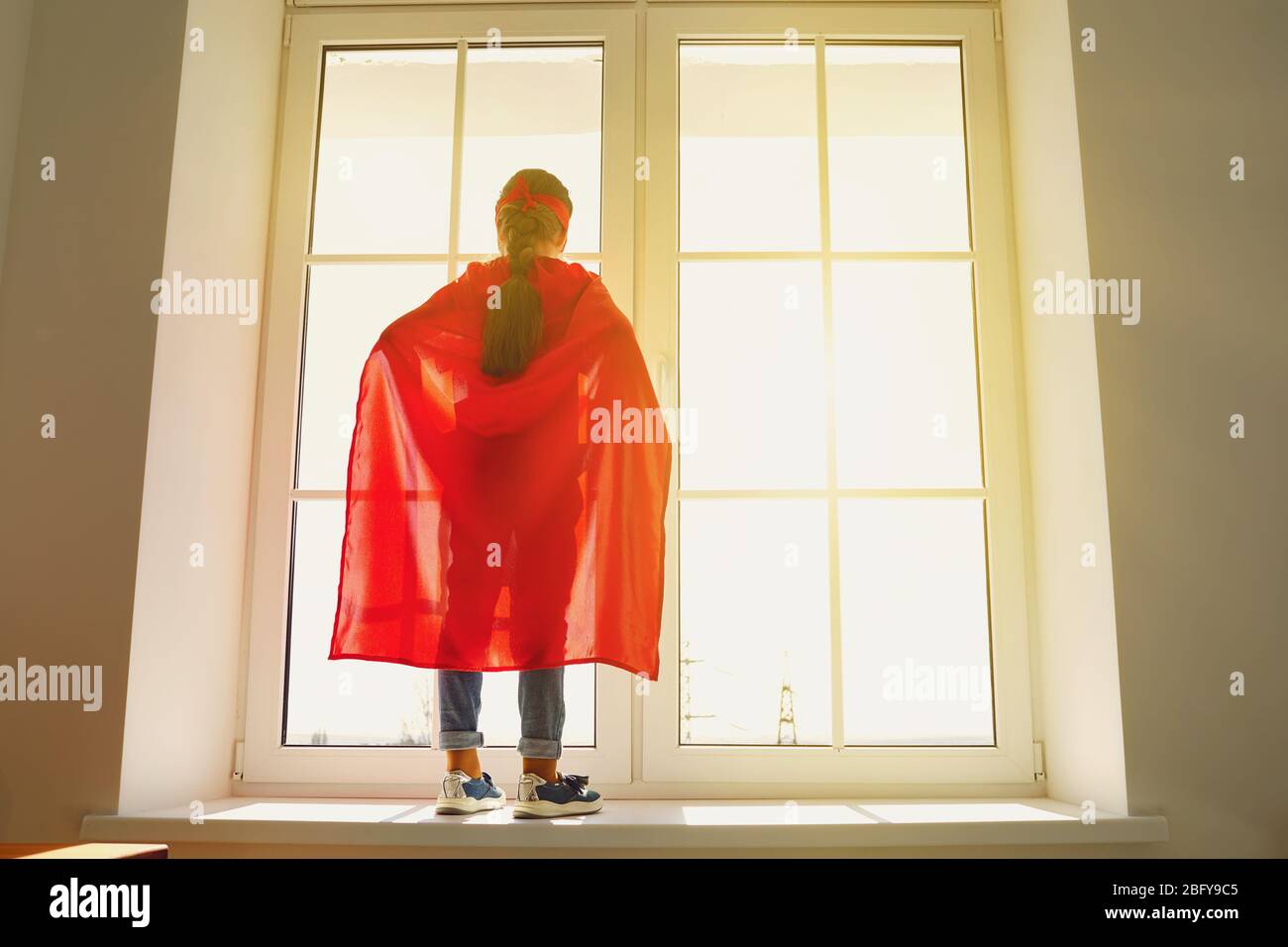 Fille enfant dans un costume de super-héros debout sur l'arrière-plan d'une fenêtre avec lumière du soleil regardant des rêves à la fenêtre Banque D'Images