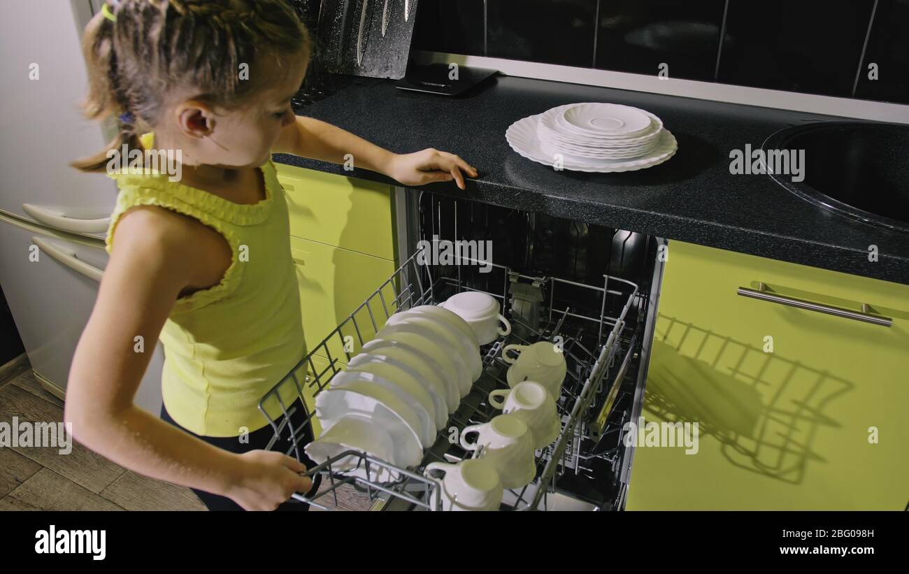 Une fille intelligente qui apprend à utiliser le lave-vaisselle. Appareils de cuisine modernes et élégants en noir vert. L'enfant met des plats propres. Banque D'Images