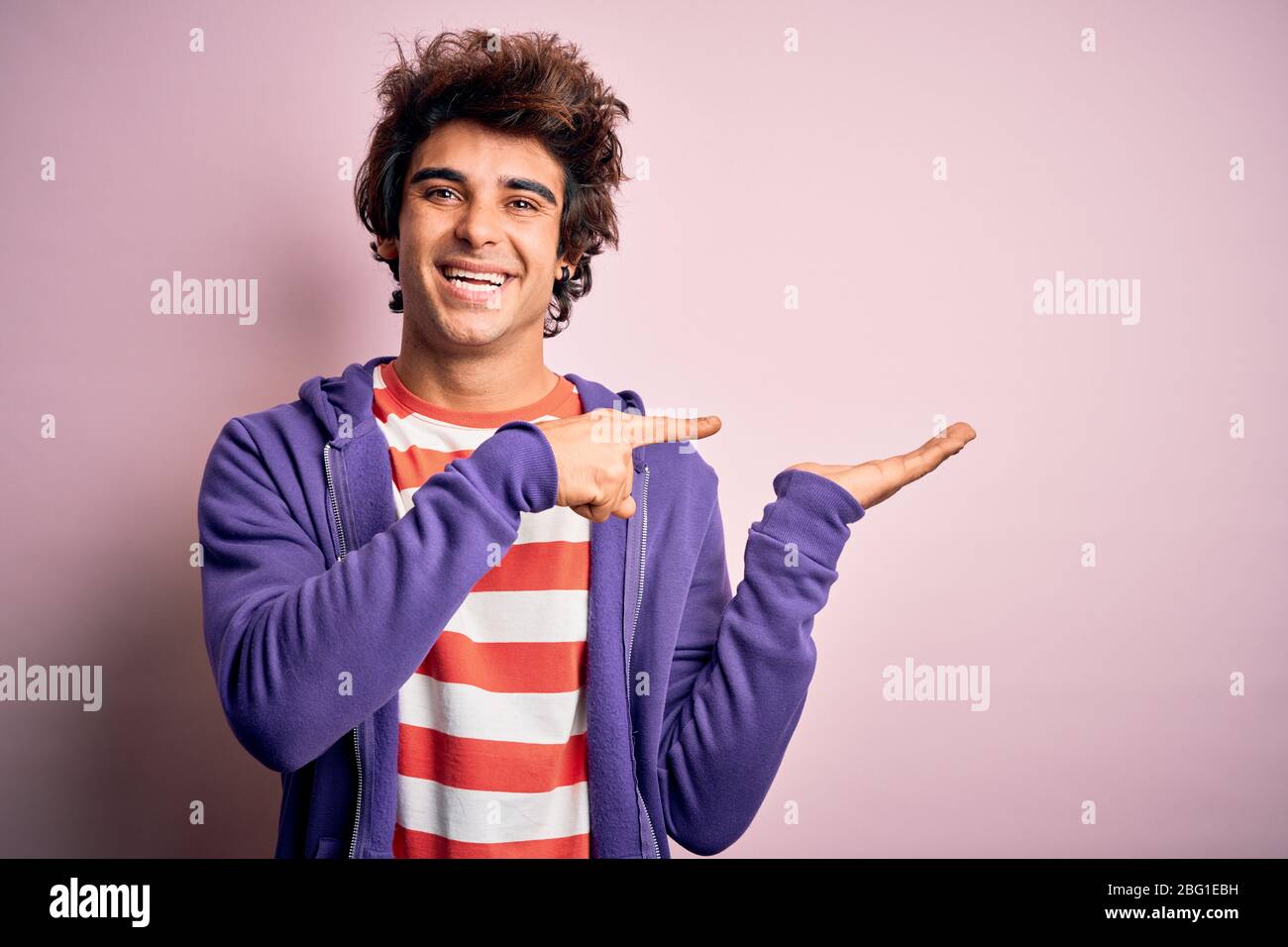 Jeune homme portant un t-shirt rayé et un sueur violet sur un fond rose isolé étonné et souriant à l'appareil photo tout en présentant avec la main et p Banque D'Images