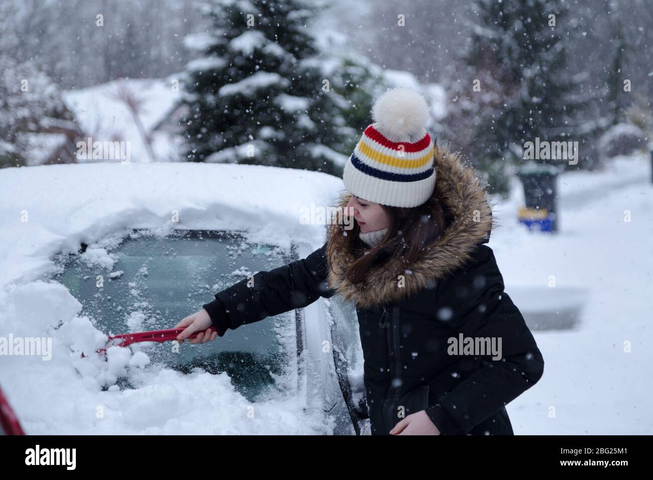 Jeune femme brunette séduisante qui déneige une voiture avec un grattoir à glace en hiver. Banque D'Images