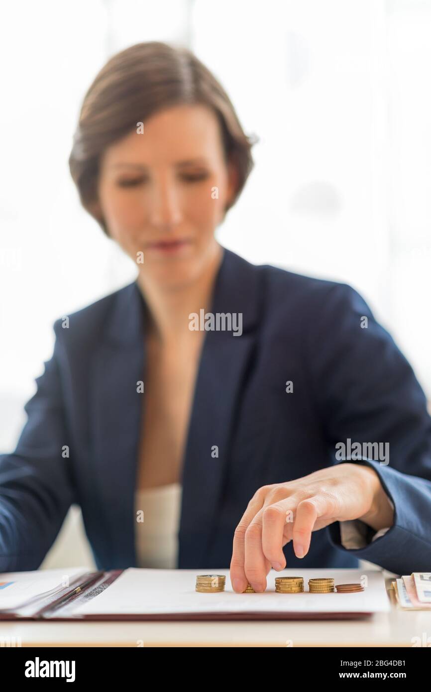 Femme d'affaires assise à son bureau équilibrant les livres comptant des piles de pièces comme elle fait la comptabilité d'affaires avec l'accent sur sa main et le m Banque D'Images