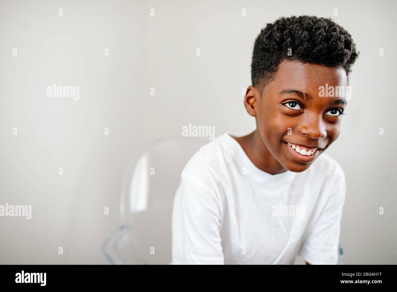 Jeune garçon noir souriant avec de grands yeux bruns chaise en acrylique clair regardant vers le haut Banque D'Images