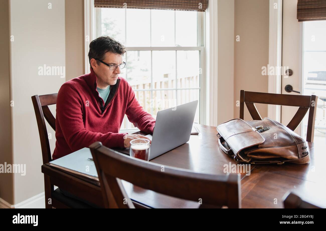 Homme dans des lunettes travaillant à la maison en utilisant un ordinateur à une table à manger. Banque D'Images