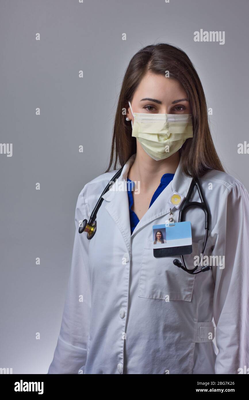 Un professionnel de la santé masqué souriant et se posant sous un masque jaune sur un fond gris. Stéthoscope, manteau blanc et EPI visibles en portrait Banque D'Images