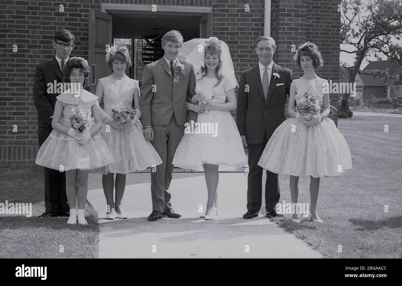 1960, historique, photo de groupe montrant des invités de mariage avec une épouse 'adolescente' dans sa robe de mariage avec voile et groom de mariée dans un costume de trois boutons de l'époque, debout ensemble à l'extérieur de l'entrée d'un bâtiment moderne d'église, Angleterre, Royaume-Uni. Banque D'Images