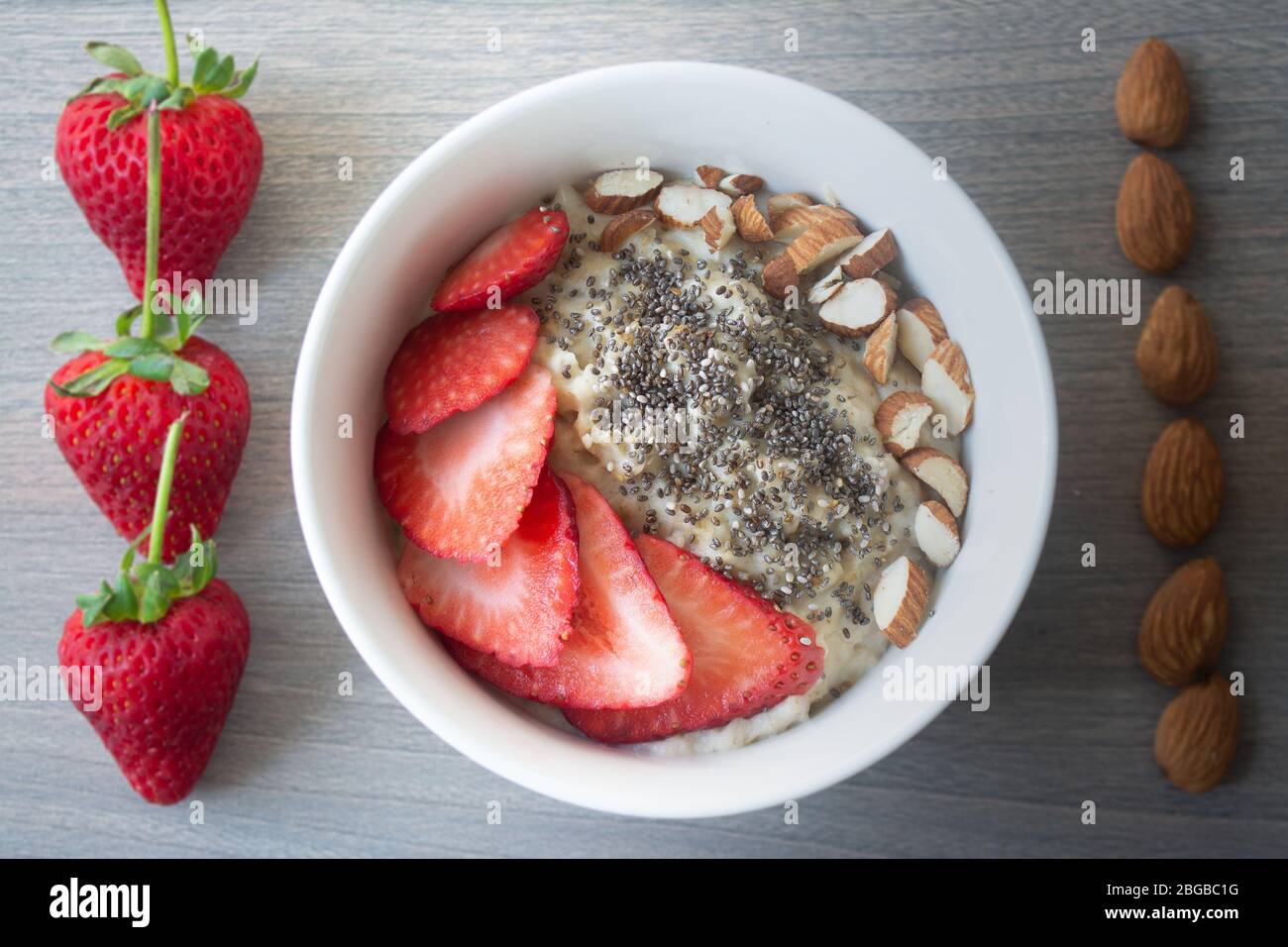 Flocons d'avoine végétalien sain avec fraises, graines de chia et amandes tranchées dans un bol blanc avec fraises et amandes entières en arrière-plan Banque D'Images