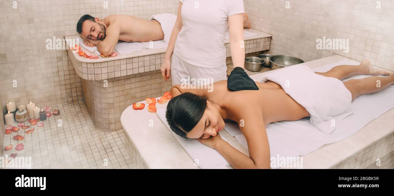 Femme de course mixte qui s'épluge la peau dans le hammam turc tandis que son petit ami bénéficie de la chaleur d'une table de massage en marbre du bain turc Banque D'Images