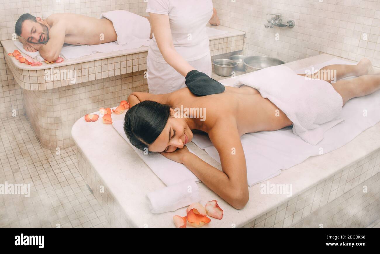 Femme de course mixte qui s'épluge la peau dans le hammam turc tandis que son petit ami bénéficie de la chaleur d'une table de massage en marbre du bain turc Banque D'Images