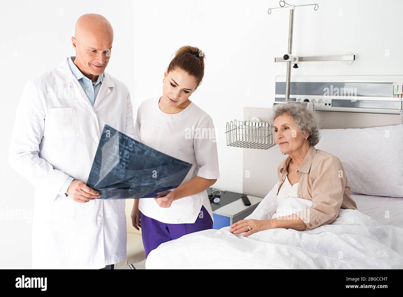 Travail d'équipe, médecin et infirmière debout dans la salle près du patient âgé, ils regardent une image d'une radiographie des vertèbres. salle hospitalière Banque D'Images