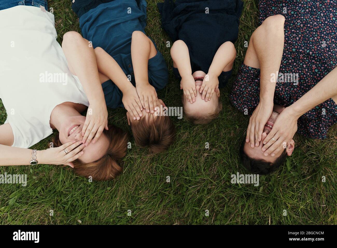 Une jeune famille de quatre membres reposant sur les yeux de couverture d'herbe avec les mains, crée des souvenirs pour une vie Banque D'Images