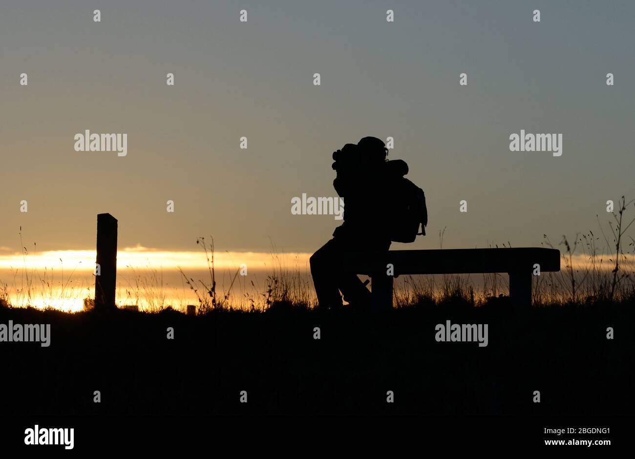 Silhouette d'un observateur d'oiseaux assis sur un banc avec des jumelles soulevées devant un coucher de soleil doré Banque D'Images