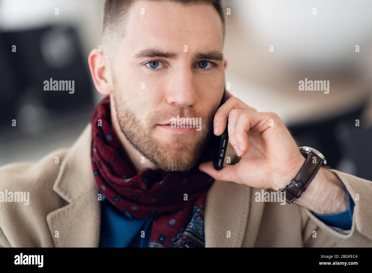 Young adult man en écharpe manteau élégant et assis à l'intérieur du terminal de l'aéroport d'attente. Il regarde l'écran du téléphone mobile cellulaire et Banque D'Images