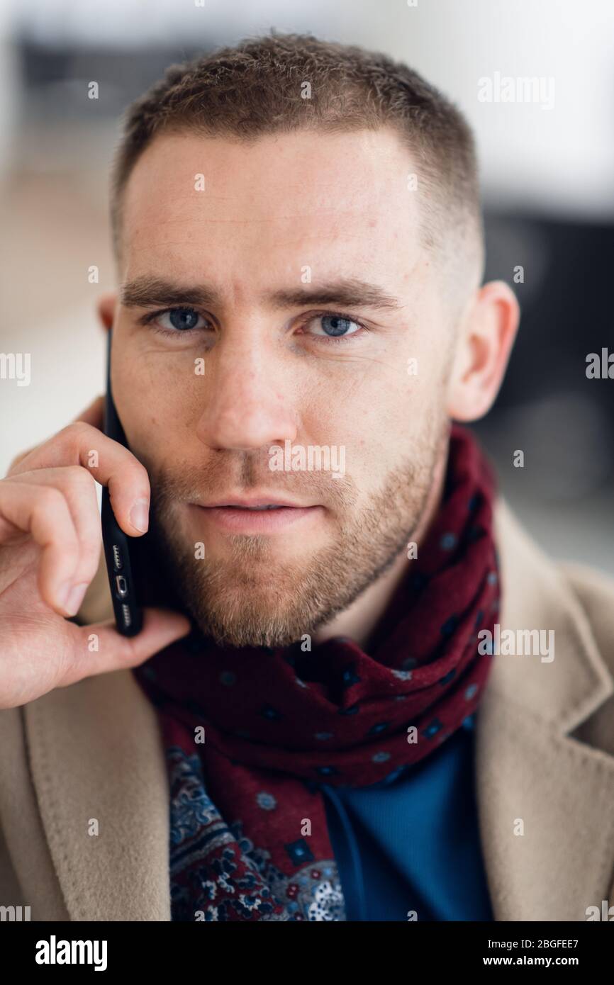 Jeune homme sérieux portant un foulard et un manteau parlant sur un téléphone portable gros plan. Banque D'Images