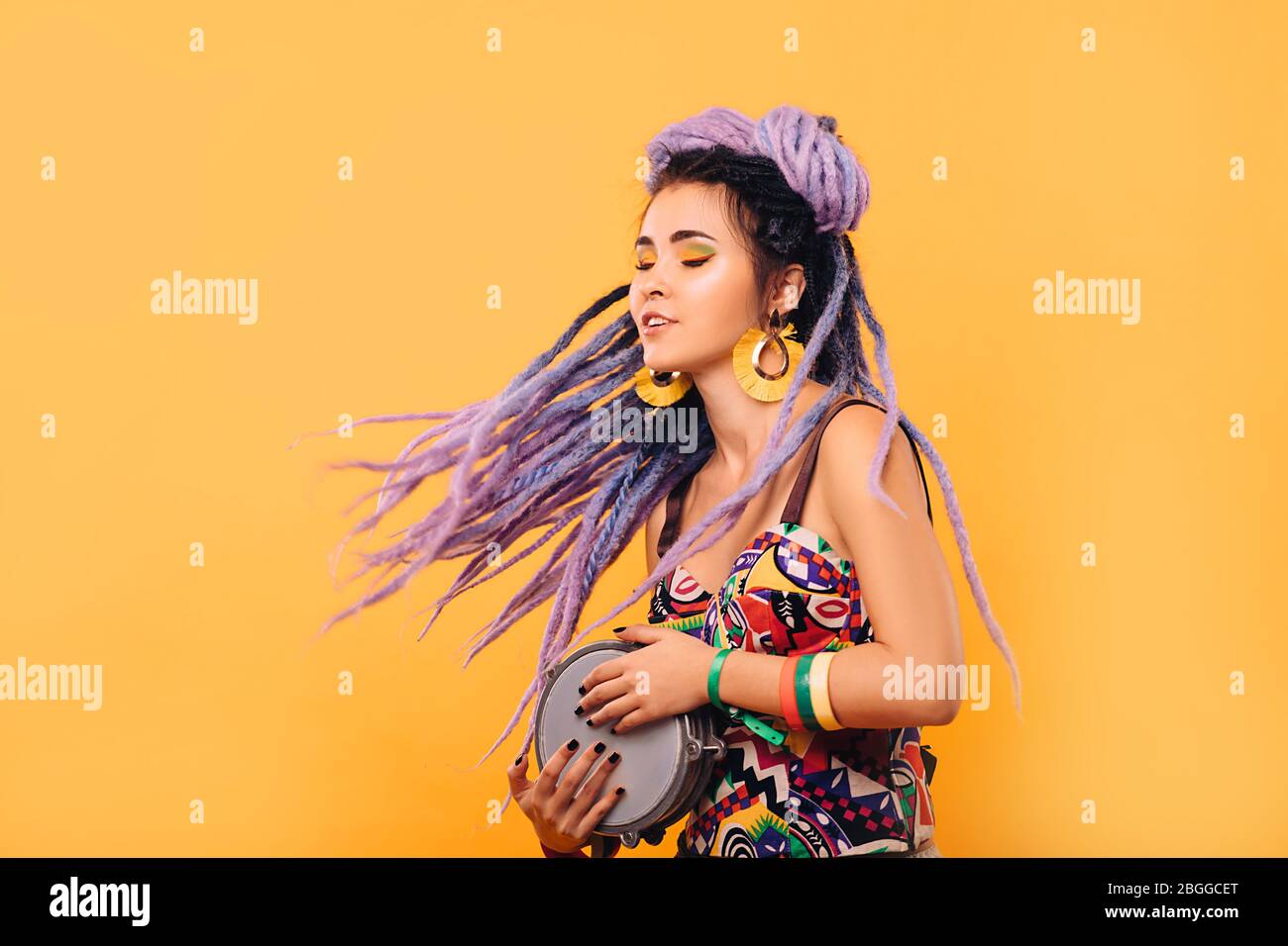 Hipster femme avec des cadenas violets et des vêtements de couleur jouant sur le mini-tambour éthique sur fond jaune Banque D'Images