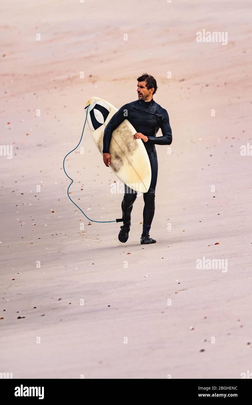 Surfeur masculin portant une combinaison, tenant planche de surf sous son bras, marchant sur la plage après la séance de surf du matin. Banque D'Images