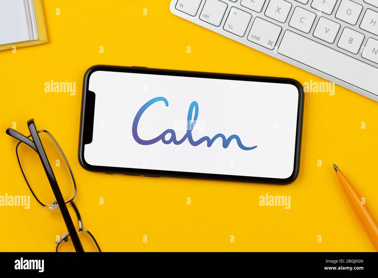 Un smartphone affichant le logo de l'application Calm repose sur un fond jaune avec un clavier, des lunettes, un stylo et un livre (usage éditorial uniquement). Banque D'Images