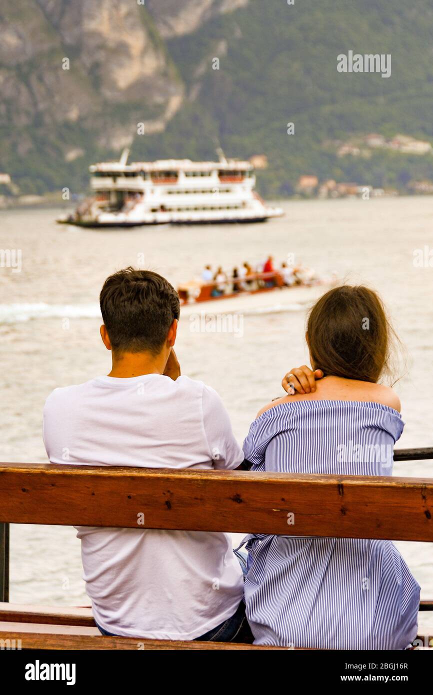 BELLAGIO, LAC DE CÔME, ITALIE - JUIN 2019: Jeune homme et femme assis sur un banc en bois donnant sur le port de Bellagio sur le lac de Côme. Banque D'Images