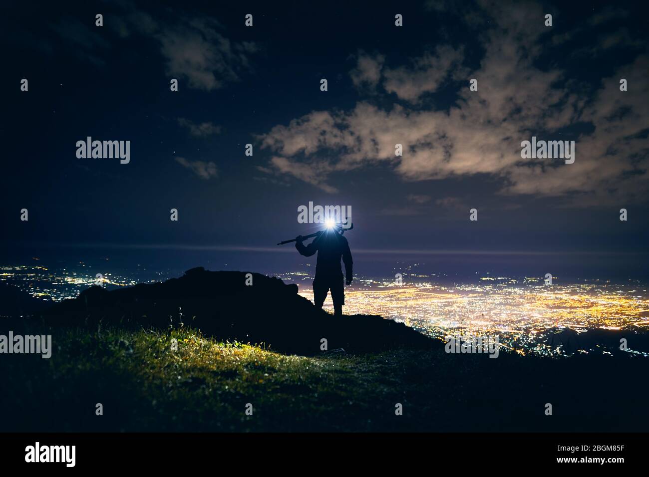 Photographe avec trépied en silhouette marcher sur les montagnes avec lumière LED lumineux contre ciel de nuit avec des étoiles et incandescent ville fond clair. Banque D'Images