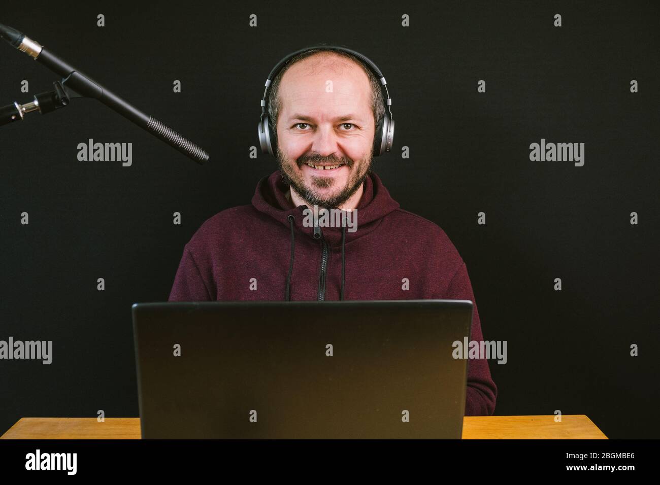 Guy dans hoodie assis derrière le bureau avec ordinateur portable et casque sur fond noir, parler au microphone, enregistrer vidéo blog Banque D'Images