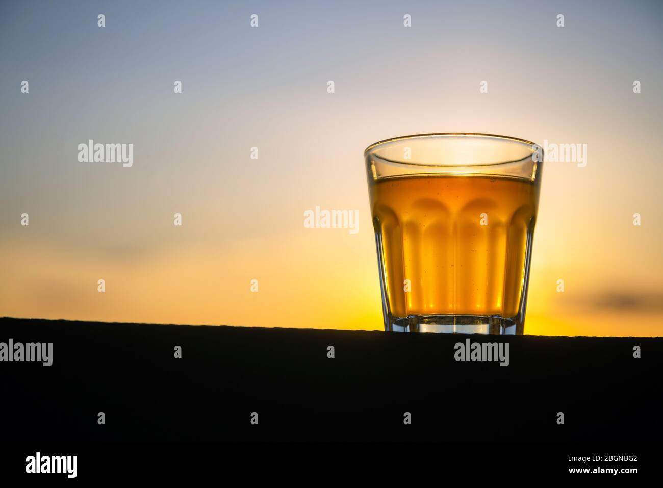 Petit verre de bière artisanale sans mousse sur une planche en bois à l'extérieur avec de l'espace pour le texte. Échantillon doré de boissons alcoolisées avec fond de coucher de soleil. Personne Banque D'Images