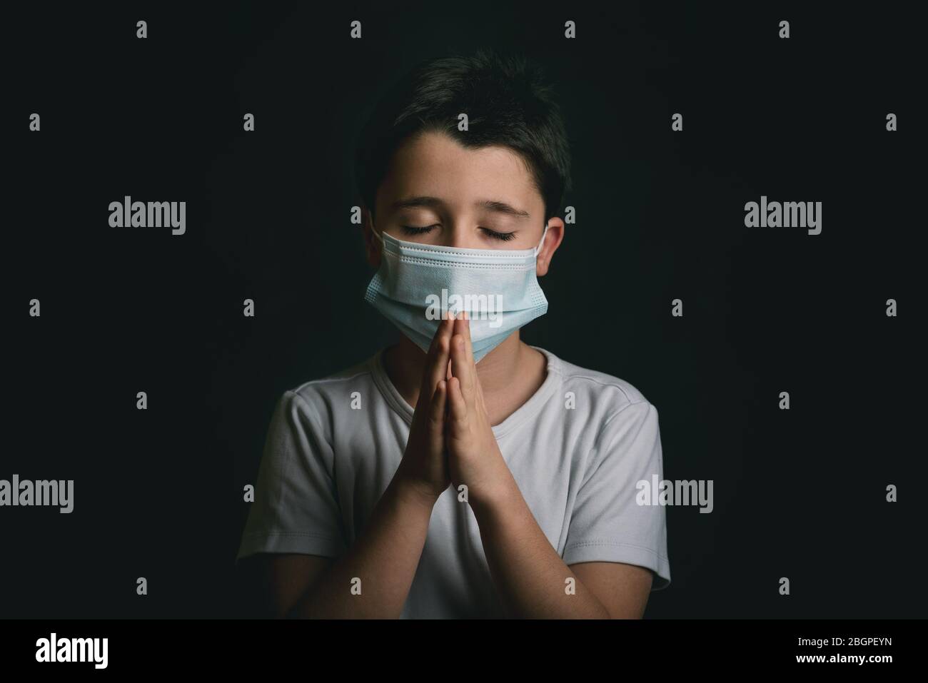 enfant portant un masque médical pour coronavirus avec des mains claquées ensemble priant sur fond noir Banque D'Images