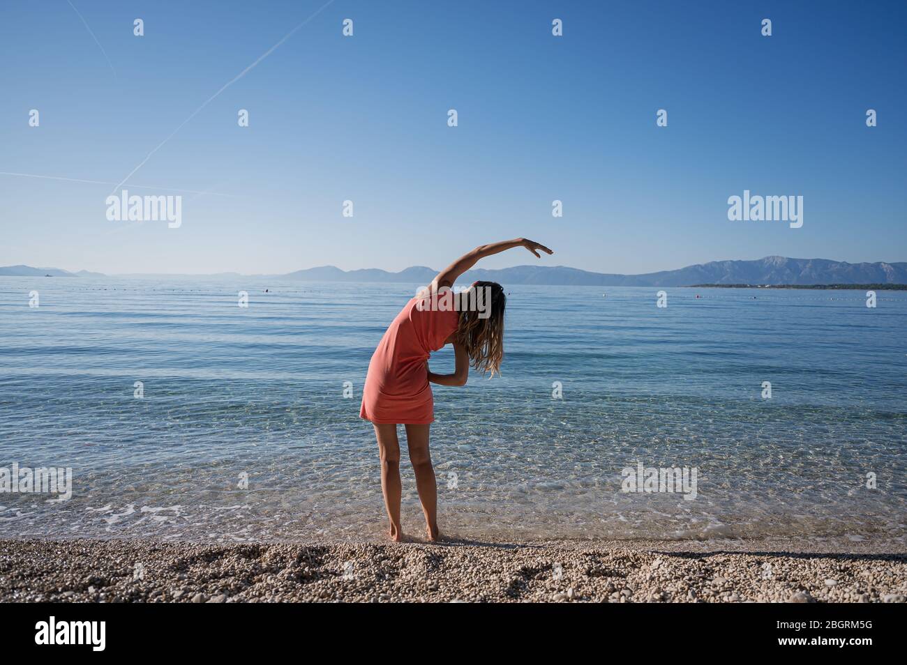 Vue de derrière une jeune femme debout cheville profondément dans l'eau de mer calme matin s'étendant avec son bras sur le côté. Banque D'Images