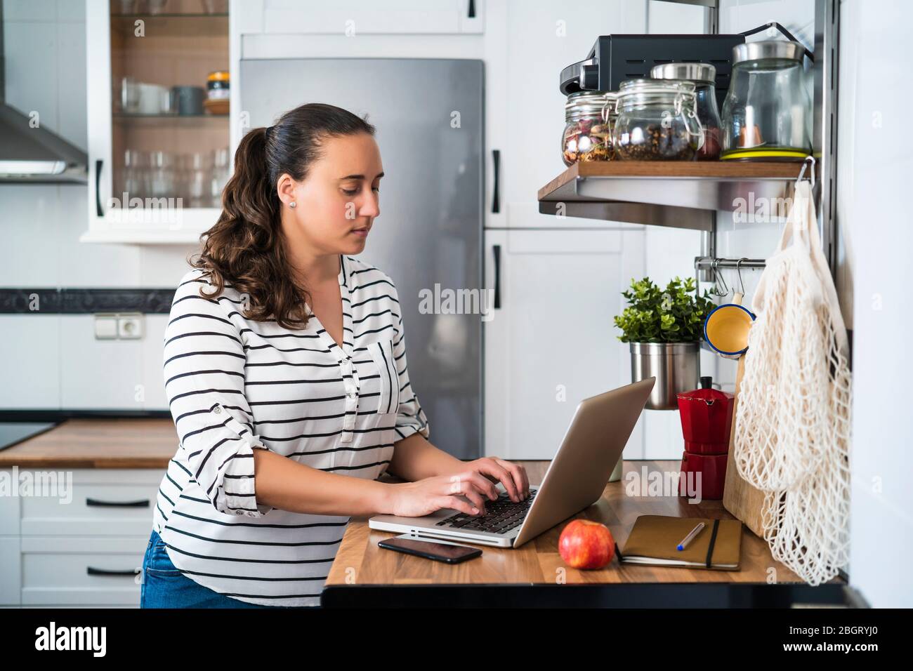 Femme souriante utilisant son ordinateur portable dans sa cuisine d'appartement. Travailler à la maison et rester à la maison. Banque D'Images