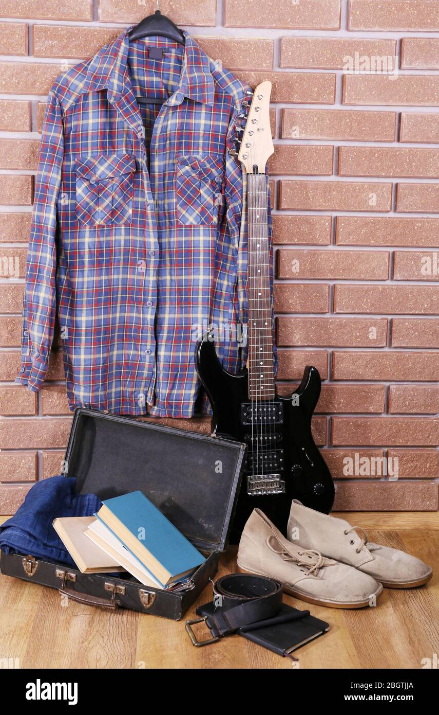 Guitare, vêtements, bottes et valise avec des livres au sol sur fond de mur de briques Banque D'Images