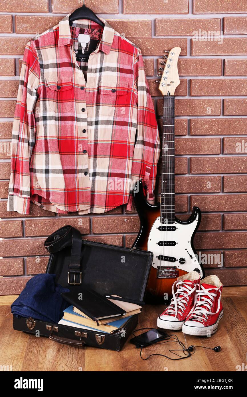 Guitare, vêtements, bottes et valise avec des livres au sol sur fond de mur de briques Banque D'Images