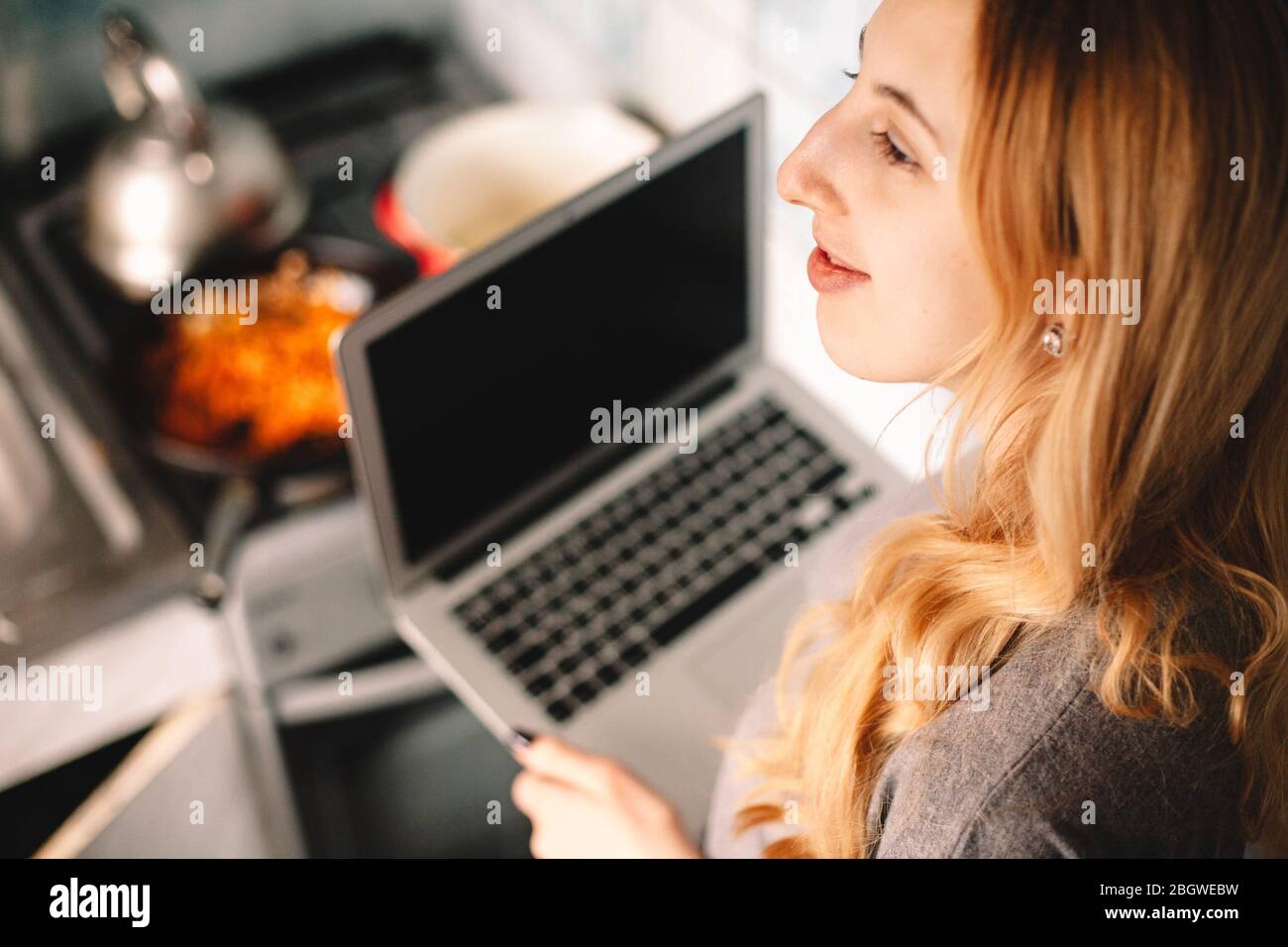 Jeune femme réfléchie tenant un ordinateur portable tout en cuisinant des aliments dans la cuisine Banque D'Images