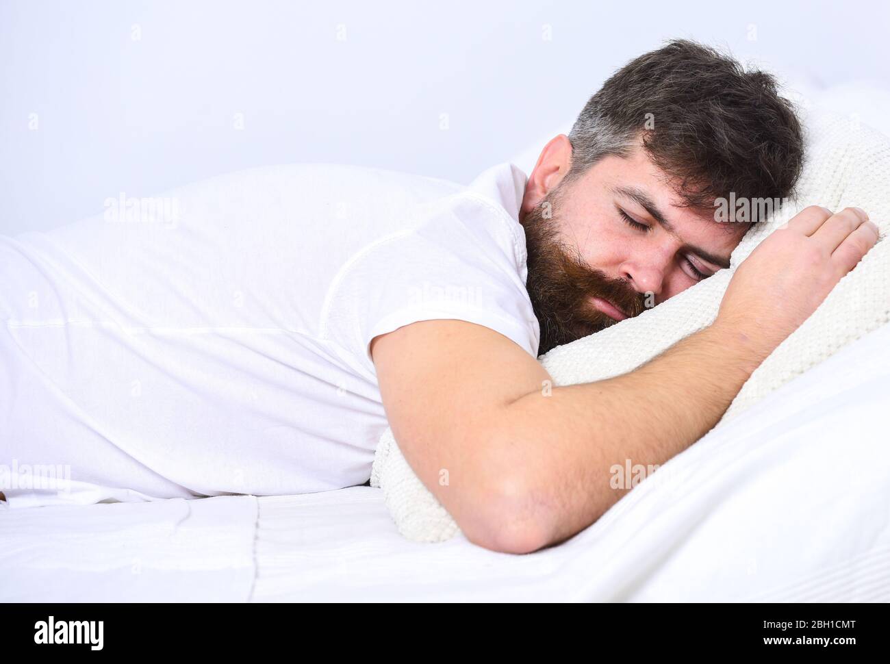 Homme en chemise posé sur le lit, mur blanc sur fond. Guy sur le visage  calme dormant sur des draps blancs et un oreiller. Concept NAP et siesta.  Macho avec barbe et