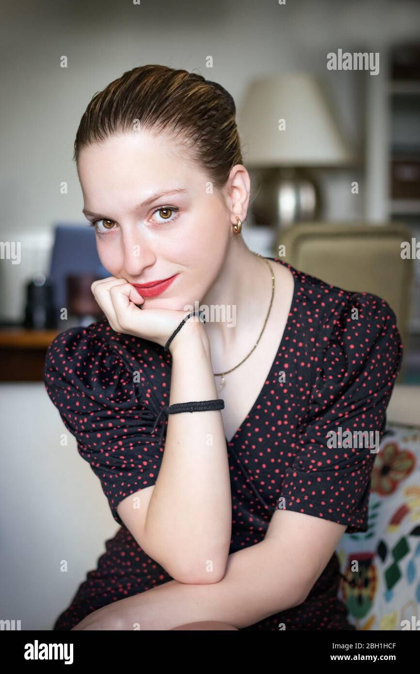 Portrait de petite adolescente avec main sur le menton, confiante, assise à la maison. Banque D'Images