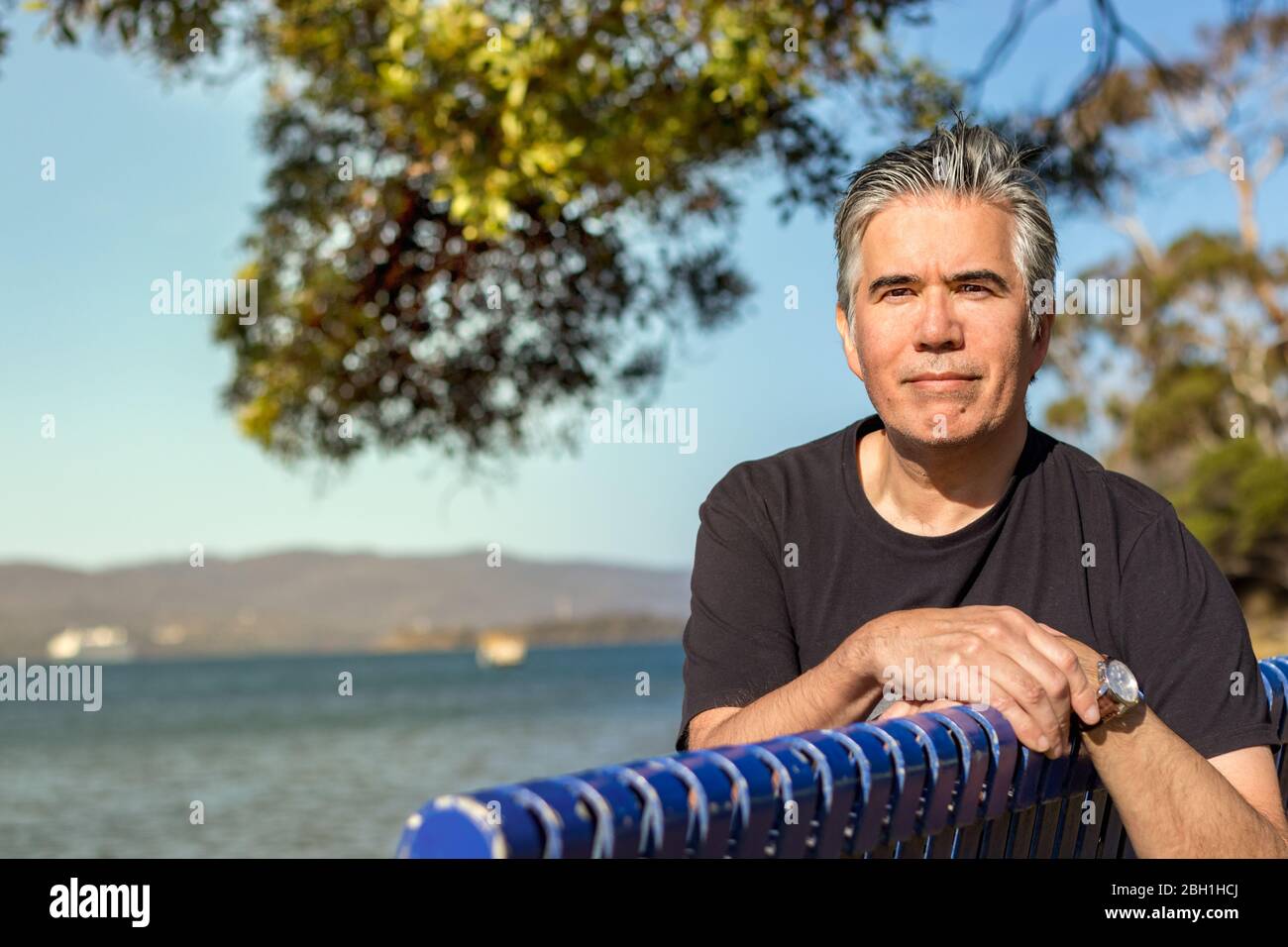 Portrait d'un homme mûr 57 ans avec un appareil photo gris à l'aspect des cheveux assis sur un banc à une plage à l'extérieur, confiant, détendu Banque D'Images