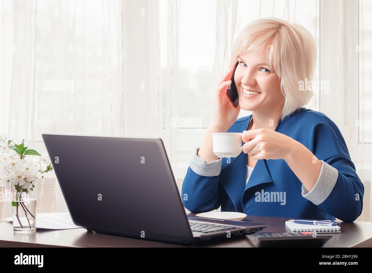 Une jolie fille souriante parle au téléphone et tient une tasse de café ou de thé dans sa main tout en travaillant à distance à partir de son bureau à la maison. Banque D'Images