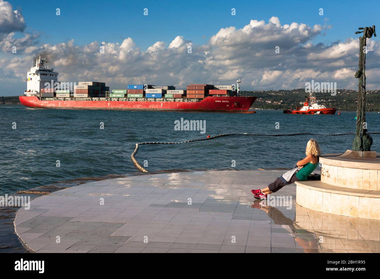 Bateau à conteneurs « King Basil » entrant dans le port de Koper, en Slovénie, surveillé par une jeune femme se reposant sur la promenade Banque D'Images